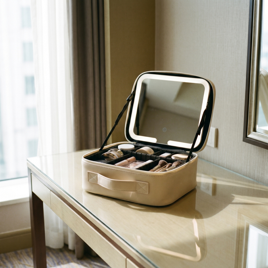 The beige travel makeup case open on a vanity, makeup sorted in its dividers, LED mirror lit